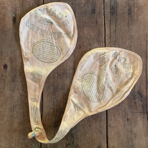 Puede incluir: Un par de alas de agua Ayvad vintage, hechas de lona blanca con costuras marrones. Las alas están conectadas por una larga correa marrón con una hebilla de metal. El texto "Ayvad Water-Wings" está impreso en negro en la lona, junto con instrucciones de uso.