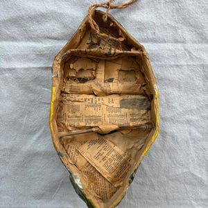 May include: Handmade paper boat with yellow trim and newspaper lining. The boat has a rustic, aged appearance with visible wear and tear. A brown twine is attached for hanging. The interior is filled with vintage newspaper clippings.