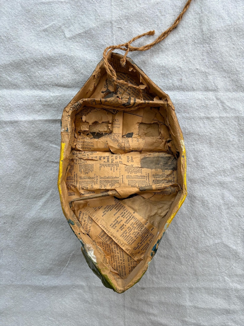 May include: Handmade paper boat with yellow trim and newspaper lining. The boat has a rustic, aged appearance with visible wear and tear. A brown twine is attached for hanging. The interior is filled with vintage newspaper clippings.