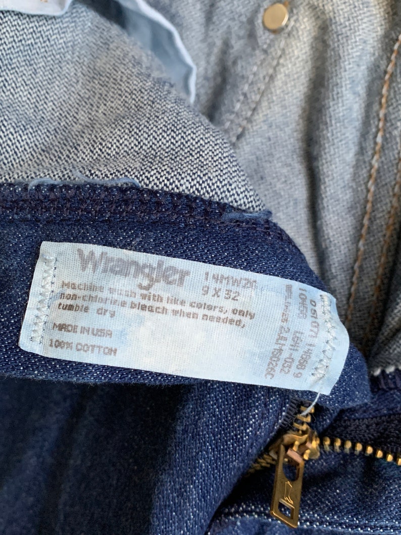 Puede incluir: Una etiqueta blanca con texto negro unida a un par de pantalones vaqueros de color azul oscuro. La etiqueta dice "Wrangler 1 MW200 9 X 32 Lavar a m&aacute;quina con colores similares, solo lej&iacute;a sin cloro cuando sea necesario, secar en secadora MADE IN USA 100% ALGOD&Oacute;N"