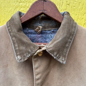 Peut inclure: Gros plan sur une veste de travail beige avec un col en velours c&ocirc;tel&eacute; et une fermeture boutonn&eacute;e. La veste est suspendue &agrave; un cintre en bois sur fond jaune. Le col est marron clair.