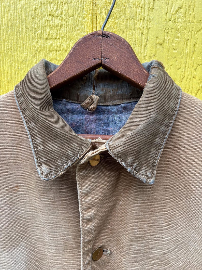 Peut inclure: Gros plan sur une veste de travail beige avec un col en velours c&ocirc;tel&eacute; et une fermeture boutonn&eacute;e. La veste est suspendue &agrave; un cintre en bois sur fond jaune. Le col est marron clair.