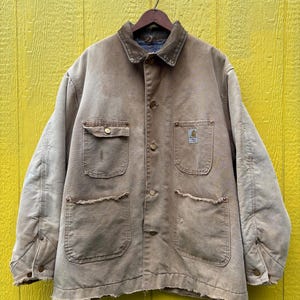 Peut inclure: Veste de travail beige avec un col en velours c&ocirc;tel&eacute; marron, quatre poches avant et des fermetures &agrave; boutons. La veste a un aspect us&eacute; et est suspendue &agrave; un cintre en bois sur un fond jaune.