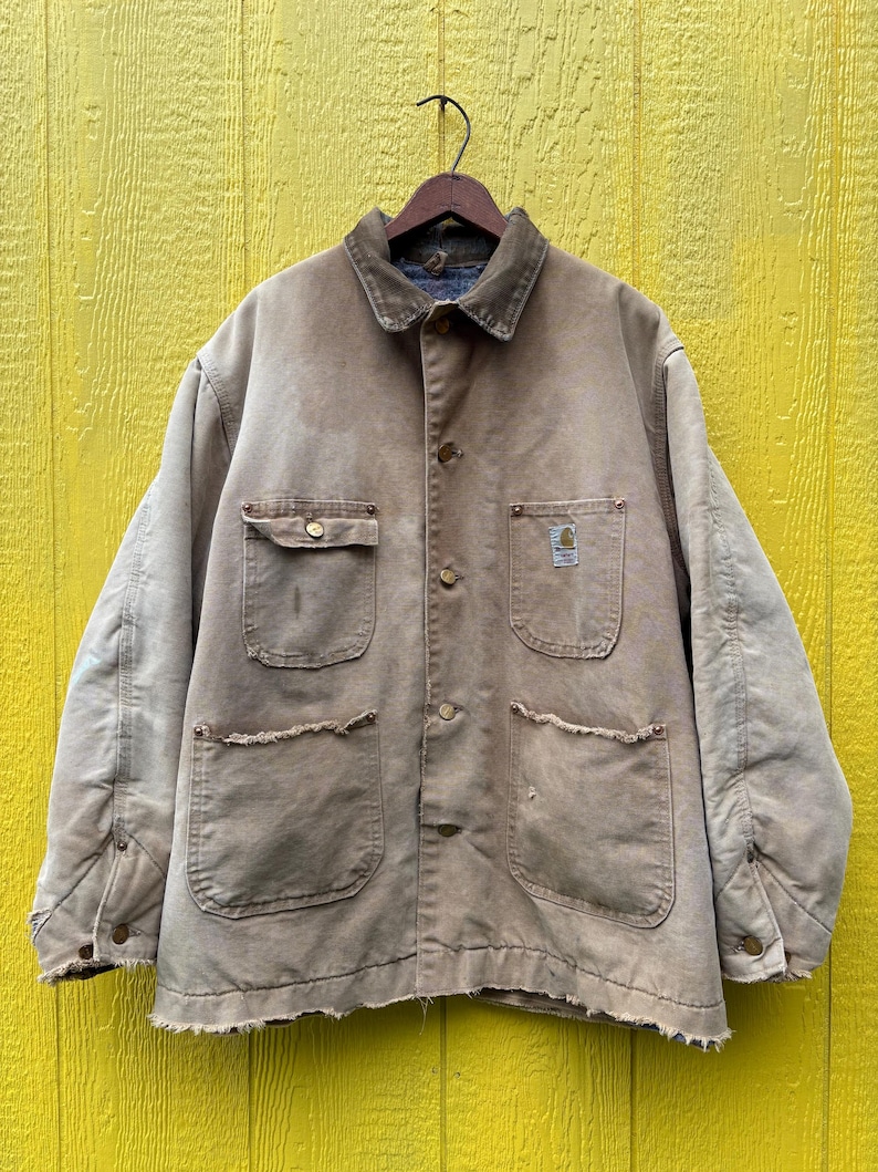Peut inclure: Veste de travail beige avec un col en velours c&ocirc;tel&eacute; marron, quatre poches avant et des fermetures &agrave; boutons. La veste a un aspect us&eacute; et est suspendue &agrave; un cintre en bois sur un fond jaune.