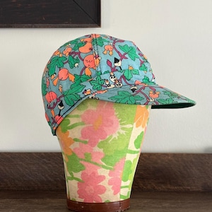 May include: A baseball cap with a colorful floral and fruit pattern on a light blue background. The cap is displayed on a floral patterned hat stand. The brim of the cap is green.
