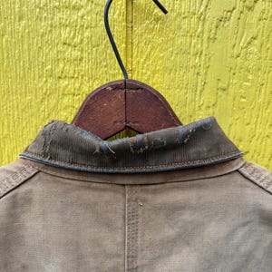 Peut inclure: Gros plan d'une veste de travail beige suspendue &agrave; un cintre en bois sur un fond jaune. La veste a un col us&eacute; et des d&eacute;tails de couture visibles. Le dos de la veste est centr&eacute; dans le cadre.