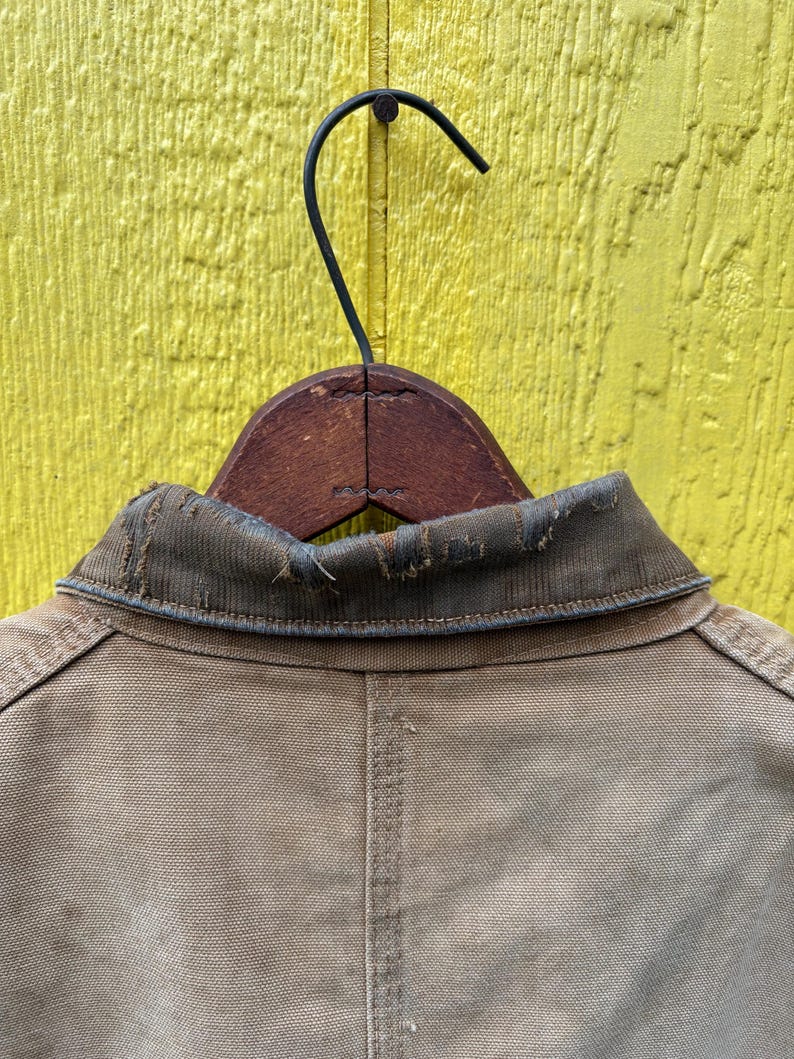 Peut inclure: Gros plan d'une veste de travail beige suspendue &agrave; un cintre en bois sur un fond jaune. La veste a un col us&eacute; et des d&eacute;tails de couture visibles. Le dos de la veste est centr&eacute; dans le cadre.