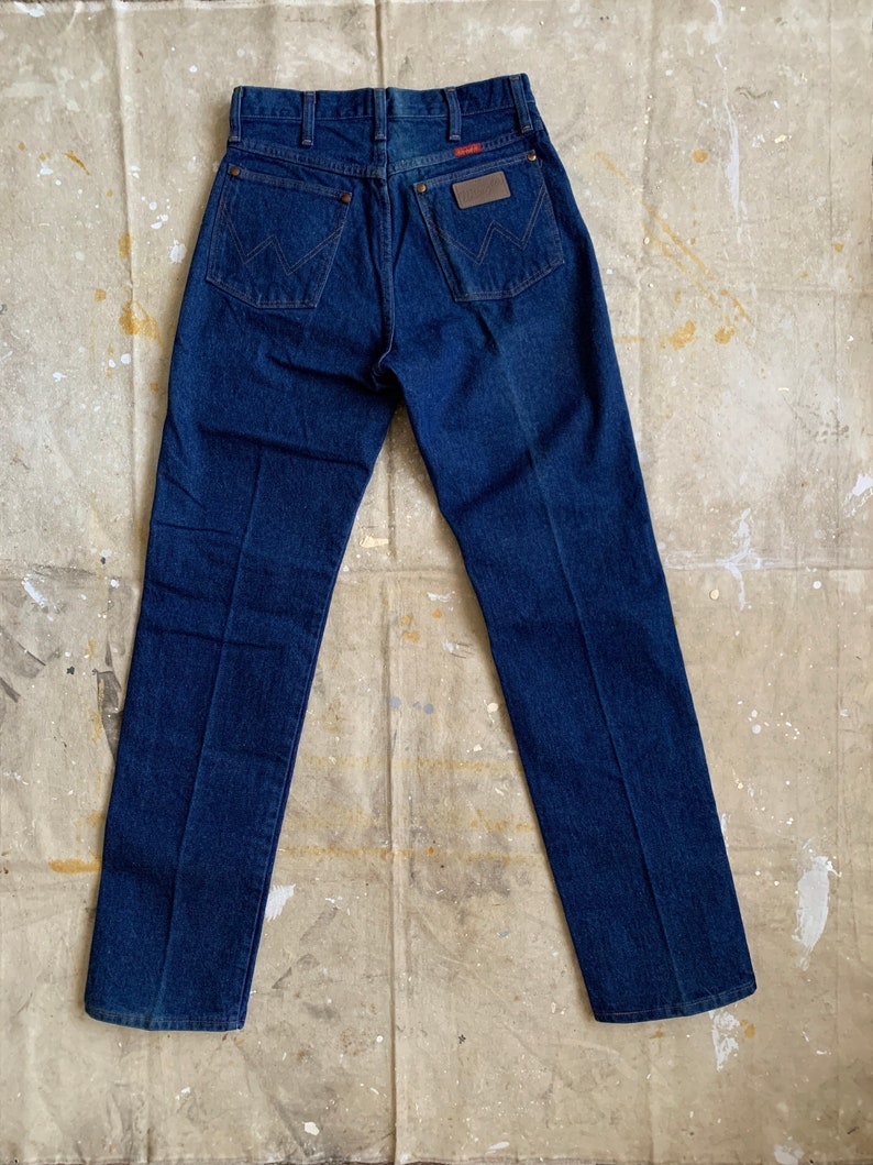Puede incluir: Un par de pantalones vaqueros de mezclilla azul oscuro con un parche de cuero en el bolsillo trasero que dice "Wrangler".