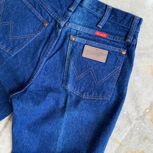 Puede incluir: Un par de pantalones vaqueros de mezclilla azul oscuro con un parche de cuero marr&oacute;n Wrangler en el bolsillo trasero.