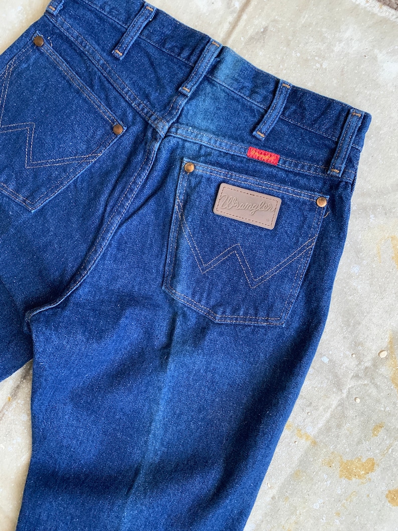 Puede incluir: Un par de pantalones vaqueros de mezclilla azul oscuro con un parche de cuero marr&oacute;n Wrangler en el bolsillo trasero.
