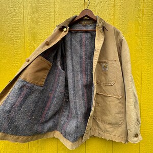 Peut inclure: Veste de travail beige avec un col en velours c&ocirc;tel&eacute; marron, suspendue &agrave; un cintre en bois. La veste a plusieurs poches et une doublure int&eacute;rieure grise et ray&eacute;e de rouge. Le logo Carhartt est visible.