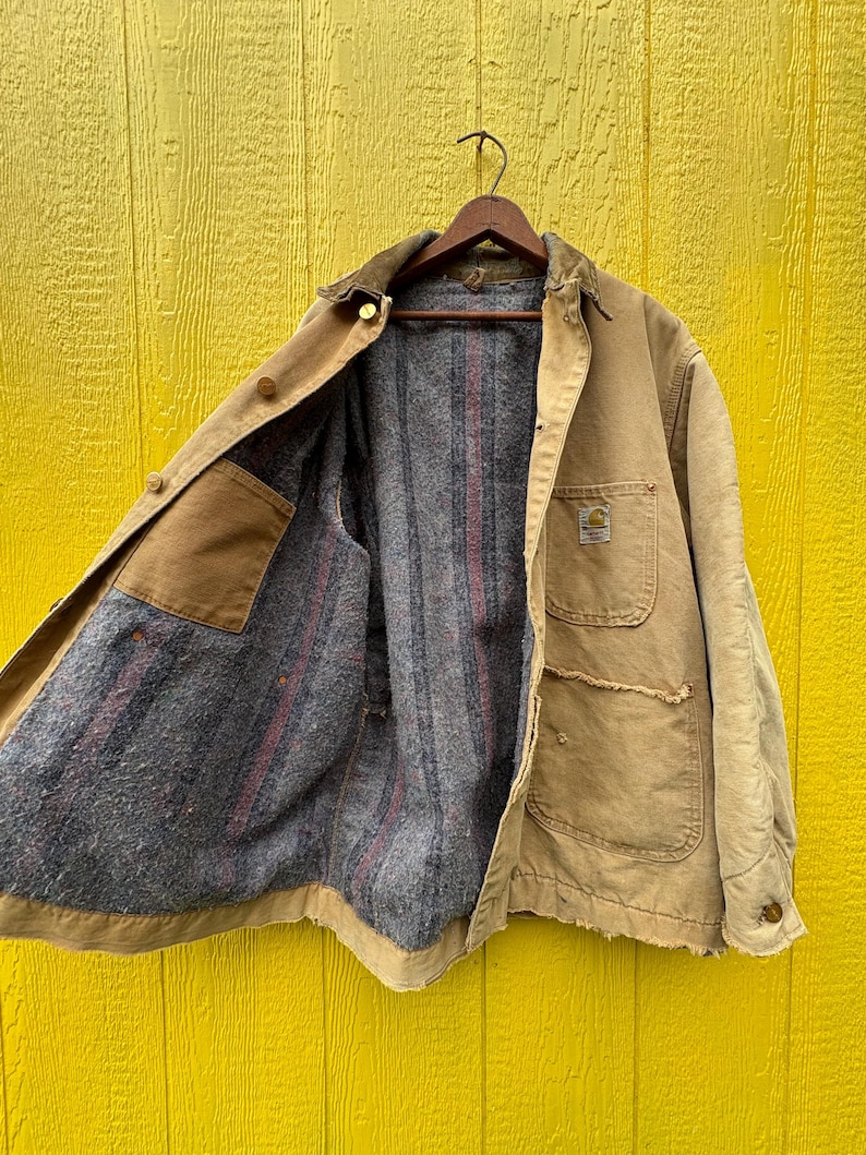 Peut inclure: Veste de travail beige avec un col en velours c&ocirc;tel&eacute; marron, suspendue &agrave; un cintre en bois. La veste a plusieurs poches et une doublure int&eacute;rieure grise et ray&eacute;e de rouge. Le logo Carhartt est visible.