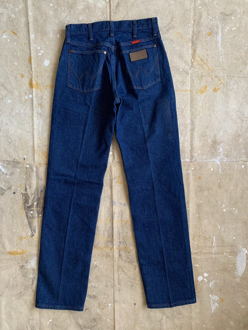 Puede incluir: Un par de pantalones vaqueros de mezclilla azul oscuro con un parche de cuero marr&oacute;n en el bolsillo trasero que dice "Wrangler".