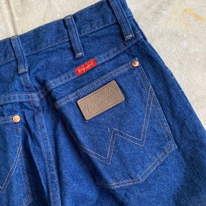 Puede incluir: Un par de pantalones vaqueros de mezclilla azul oscuro con un parche de cuero marr&oacute;n Wrangler en el bolsillo trasero.