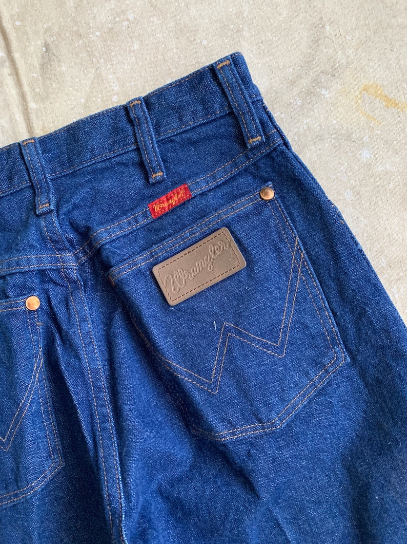 Puede incluir: Un par de pantalones vaqueros de mezclilla azul oscuro con un parche de cuero marr&oacute;n Wrangler en el bolsillo trasero.