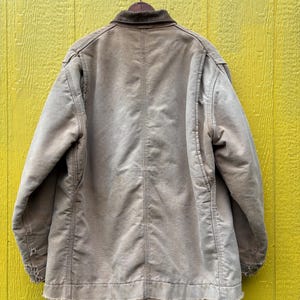 Peut inclure: Veste de travail beige avec un col en velours c&ocirc;tel&eacute; marron, suspendue &agrave; un crochet m&eacute;tallique sur un fond jaune. La veste pr&eacute;sente des signes d'usure, avec des bords effiloch&eacute;s sur les manches et l'ourlet. Le dos de la veste est visible.