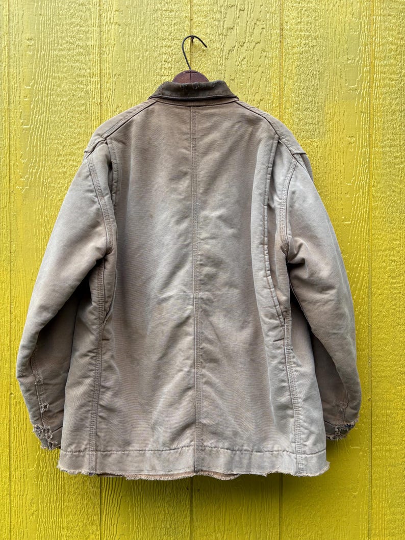Peut inclure: Veste de travail beige avec un col en velours c&ocirc;tel&eacute; marron, suspendue &agrave; un crochet m&eacute;tallique sur un fond jaune. La veste pr&eacute;sente des signes d'usure, avec des bords effiloch&eacute;s sur les manches et l'ourlet. Le dos de la veste est visible.