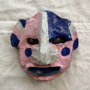 May include: A handcrafted mask with a pink face, blue and white details, and dark eyes. The mask features a smiling mouth and blue polka dots on the pink side. The nose is white, and the other side of the face is blue.