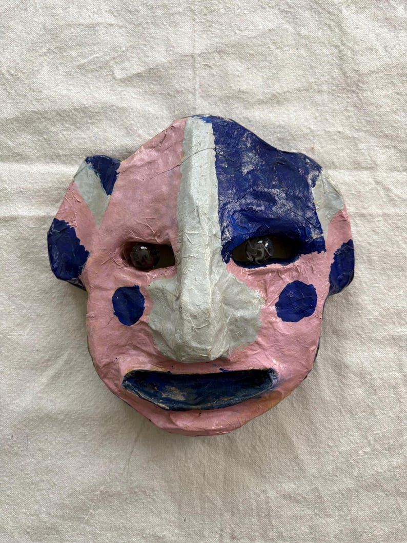 May include: A handcrafted mask with a pink face, blue and white details, and dark eyes. The mask features a smiling mouth and blue polka dots on the pink side. The nose is white, and the other side of the face is blue.