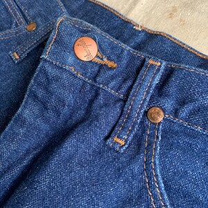 Puede incluir: Primer plano de un par de pantalones vaqueros azules de mezclilla con un bot&oacute;n de cobre y detalles de costura.