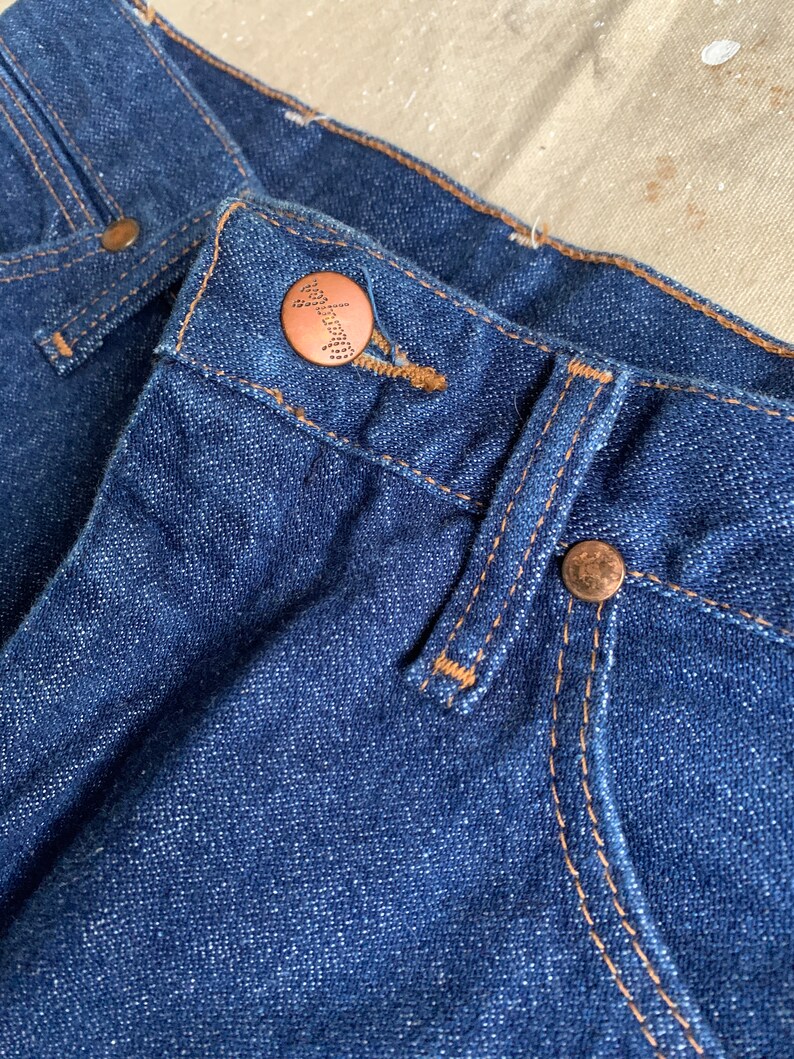 Puede incluir: Primer plano de un par de pantalones vaqueros azules de mezclilla con un bot&oacute;n de cobre y detalles de costura.