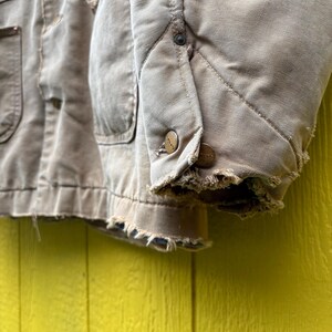 Peut inclure: Gros plan sur une veste de travail beige us&eacute;e. La veste comporte plusieurs poches, des boutons et des bords effiloch&eacute;s. Le tissu semble &ecirc;tre une toile r&eacute;sistante, avec une doublure matelass&eacute;e visible sur la manche.