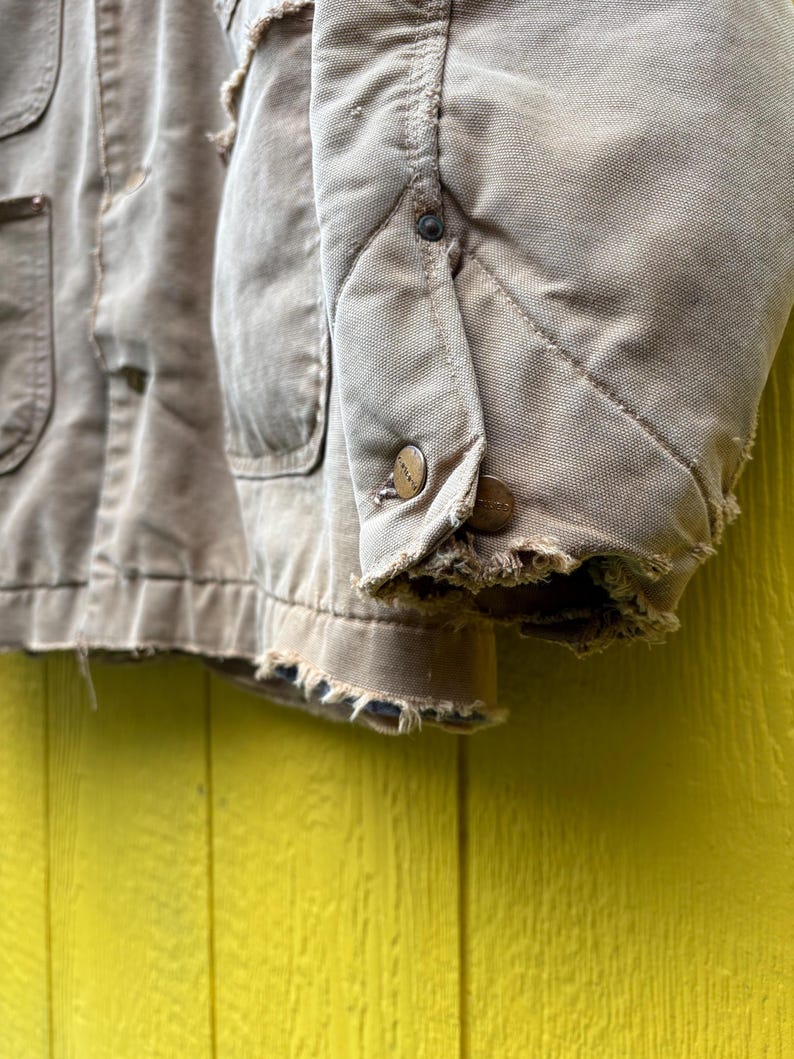 Peut inclure: Gros plan sur une veste de travail beige us&eacute;e. La veste comporte plusieurs poches, des boutons et des bords effiloch&eacute;s. Le tissu semble &ecirc;tre une toile r&eacute;sistante, avec une doublure matelass&eacute;e visible sur la manche.
