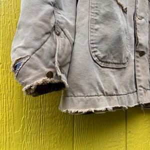 Peut inclure: Gros plan sur une veste de travail beige us&eacute;e. La veste pr&eacute;sente plusieurs poches, des boutons et un ourlet effiloch&eacute;. La manche matelass&eacute;e montre des signes d'utilisation, avec des bords effiloch&eacute;s et un aspect us&eacute;. La veste est pos&eacute;e sur un fond jaune.