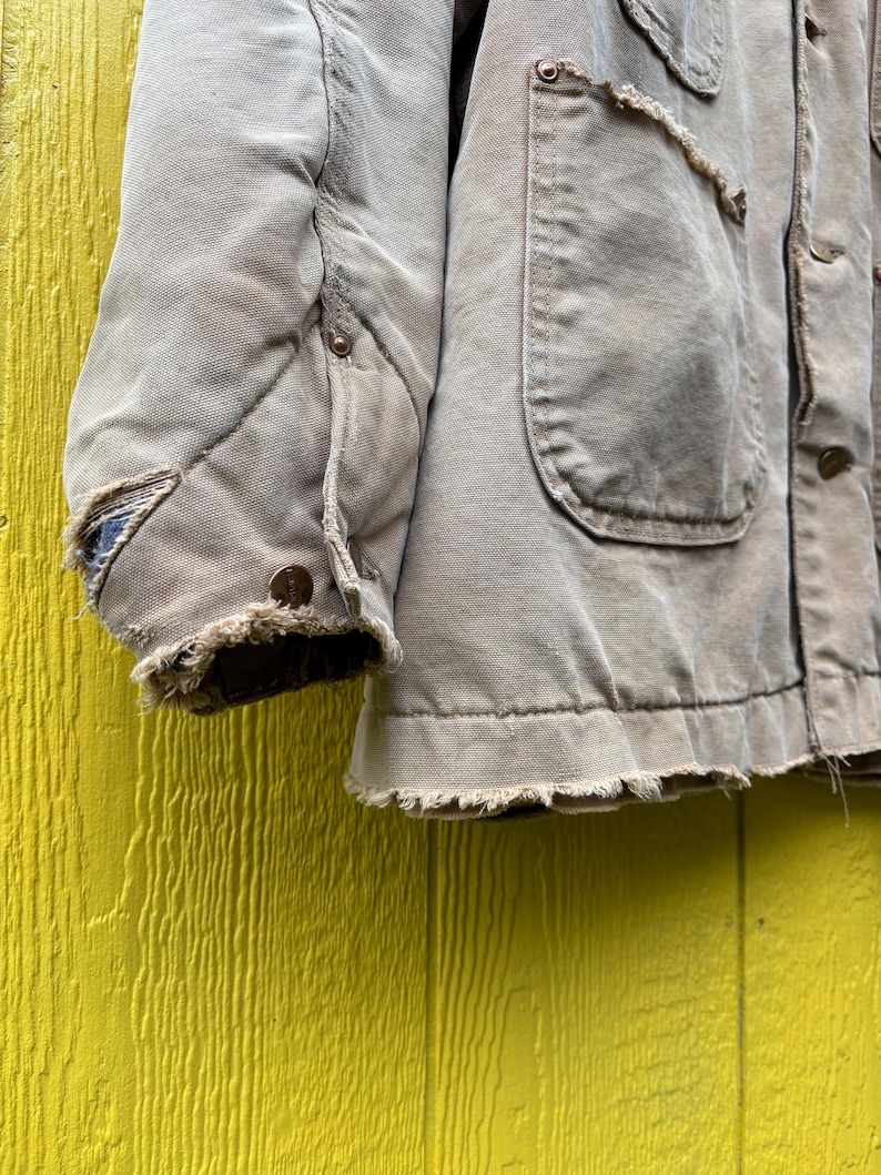 Peut inclure: Gros plan sur une veste de travail beige us&eacute;e. La veste pr&eacute;sente plusieurs poches, des boutons et un ourlet effiloch&eacute;. La manche matelass&eacute;e montre des signes d'utilisation, avec des bords effiloch&eacute;s et un aspect us&eacute;. La veste est pos&eacute;e sur un fond jaune.