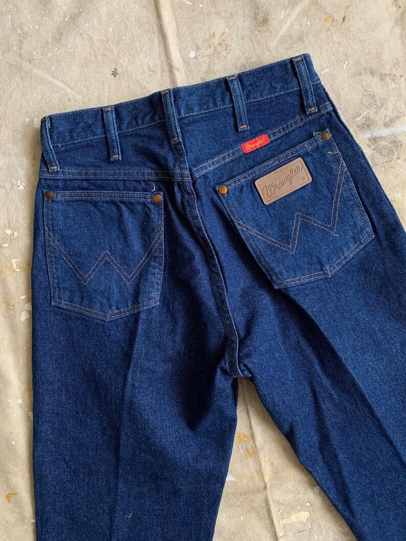Puede incluir: Un par de pantalones vaqueros de mezclilla azul oscuro con un parche de cuero marr&oacute;n Wrangler en el bolsillo trasero.