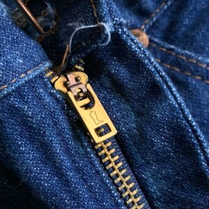 Puede incluir: Primer plano de una cremallera de mezclilla azul con un tirador de cremallera dorado. El tirador de la cremallera tiene un peque&ntilde;o dise&ntilde;o de bota.
