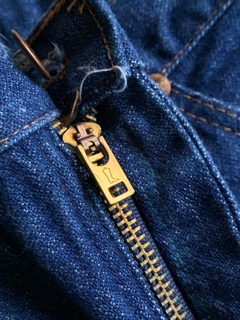 Puede incluir: Primer plano de una cremallera de mezclilla azul con un tirador de cremallera dorado. El tirador de la cremallera tiene un peque&ntilde;o dise&ntilde;o de bota.