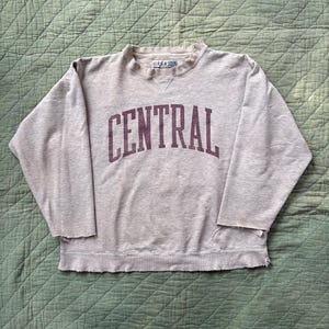 May include: A light grey sweatshirt featuring the word "CENTRAL" in maroon arched lettering. The crew neck sweatshirt has long sleeves with distressed edges. The brand "GEAR" is visible on the collar. The garment is laid on a quilted surface.