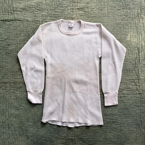 Peut inclure: T-shirt à manches longues en tricot gaufré blanc cassé, avec un col rond et des poignets côtelés. Le t-shirt est posé à plat sur une surface verte texturée. Le tissu présente un motif quadrillé.