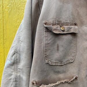 Peut inclure: Gros plan sur une veste de travail beige us&eacute;e. La veste comporte deux poches avant, dont une avec une fermeture &agrave; bouton, et des bords effiloch&eacute;s. Le tissu montre des signes d'usure et de taches, avec des coutures et des rivets m&eacute;talliques visibles.