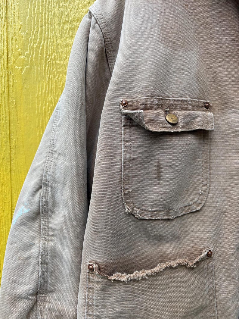 Peut inclure: Gros plan sur une veste de travail beige us&eacute;e. La veste comporte deux poches avant, dont une avec une fermeture &agrave; bouton, et des bords effiloch&eacute;s. Le tissu montre des signes d'usure et de taches, avec des coutures et des rivets m&eacute;talliques visibles.
