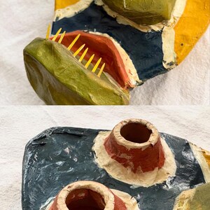 May include: A handcrafted paper mache mask with a vibrant design. It features a green, textured head, a red mouth with yellow teeth, and a blue, yellow, and white patterned base. Includes two red and white cone-shaped elements.