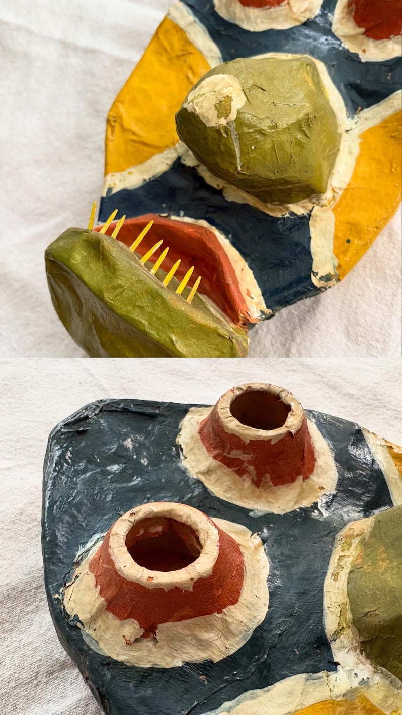 May include: A handcrafted paper mache mask with a vibrant design. It features a green, textured head, a red mouth with yellow teeth, and a blue, yellow, and white patterned base. Includes two red and white cone-shaped elements.