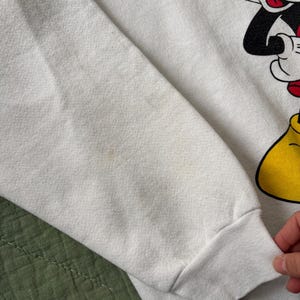 May include: White sweatshirt featuring a large graphic of Mickey Mouse. The sleeve is visible, showing the cuff and part of the arm. The design includes black, red, and yellow details. The fabric shows some discoloration.