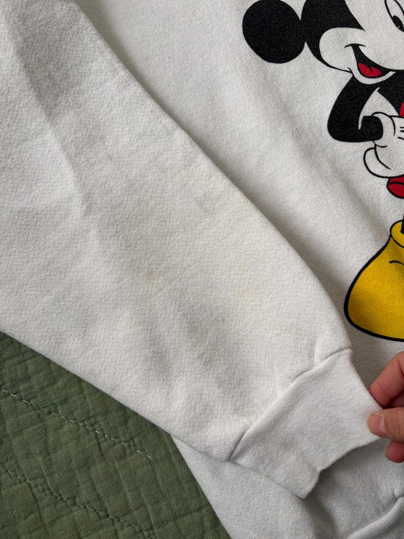 May include: White sweatshirt featuring a large graphic of Mickey Mouse. The sleeve is visible, showing the cuff and part of the arm. The design includes black, red, and yellow details. The fabric shows some discoloration.