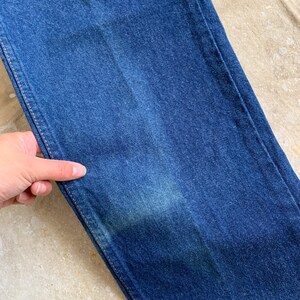 Puede incluir: Primer plano de un par de pantalones vaqueros de mezclilla azul oscuro. La tela tiene un ligero brillo y una superficie texturizada.