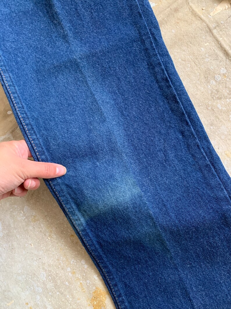 Puede incluir: Primer plano de un par de pantalones vaqueros de mezclilla azul oscuro. La tela tiene un ligero brillo y una superficie texturizada.