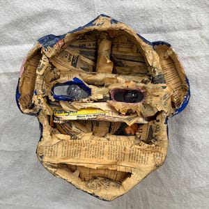 May include: A handmade paper mache mask in the shape of a face. The mask is constructed from newspaper and painted with blue and pink accents. The eyes are made of reflective material.