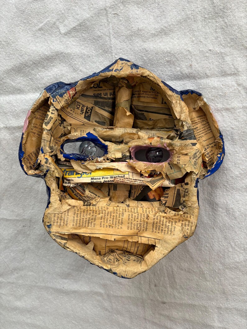 May include: A handmade paper mache mask in the shape of a face. The mask is constructed from newspaper and painted with blue and pink accents. The eyes are made of reflective material.