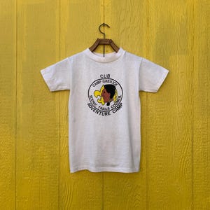 Könnte beinhalten: Weißes T-Shirt mit einem Vintage-Logo eines Pfadfinderlagers. Das Logo zeigt eine indianische Frau in einem Kopfschmuck, eine Lilie und den Text "Camp Greilick, Scenic Trails Council, Adventure Camp, Cub".