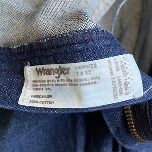 Puede incluir: Una etiqueta de mezclilla Wrangler azul con texto blanco. La etiqueta dice "Wrangler 14MWZG 7x32" e incluye instrucciones de cuidado: "Lavar a m&aacute;quina con colores similares, solo blanqueador sin cloro cuando sea necesario, secar en secadora. Hecho en EE. UU. 100% algod&oacute;n."