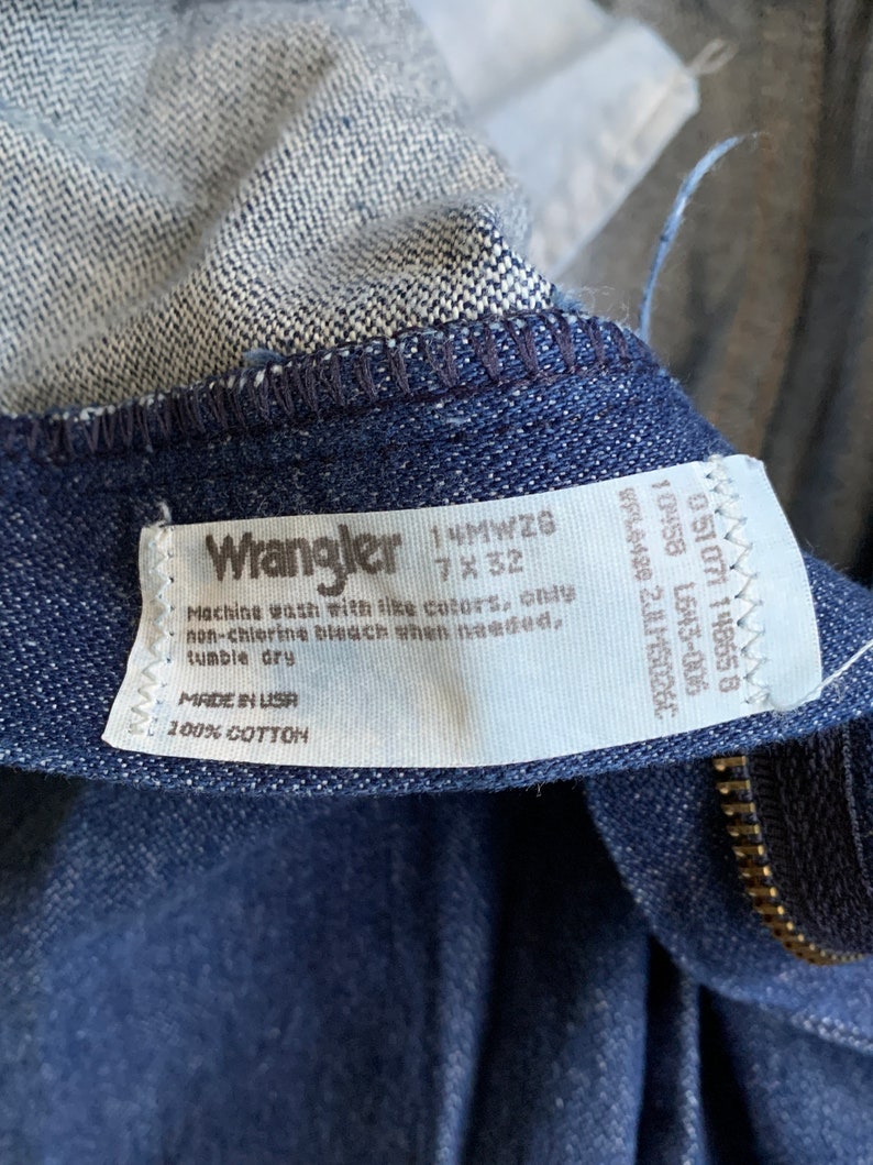 Puede incluir: Una etiqueta de mezclilla Wrangler azul con texto blanco. La etiqueta dice "Wrangler 14MWZG 7x32" e incluye instrucciones de cuidado: "Lavar a m&aacute;quina con colores similares, solo blanqueador sin cloro cuando sea necesario, secar en secadora. Hecho en EE. UU. 100% algod&oacute;n."