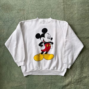May include: White crew neck sweatshirt featuring a graphic of Mickey Mouse. Mickey is depicted with black ears, a black face, red shorts, yellow shoes, and white gloves. The sweatshirt is made of a soft material.