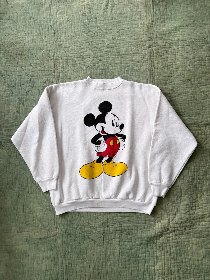 May include: White crew neck sweatshirt featuring a graphic of Mickey Mouse. Mickey is depicted with black ears, a black face, red shorts, yellow shoes, and white gloves. The sweatshirt is made of a soft material.