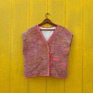 May include: A pink and multi-colored woven vest with a V-neck and two small pockets. The vest has a pink trim and is hanging on a wooden hanger against a yellow background.